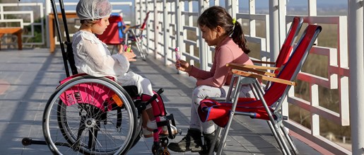 A child using a wheelchair chatting to a child in a deck chair sitting on a pier